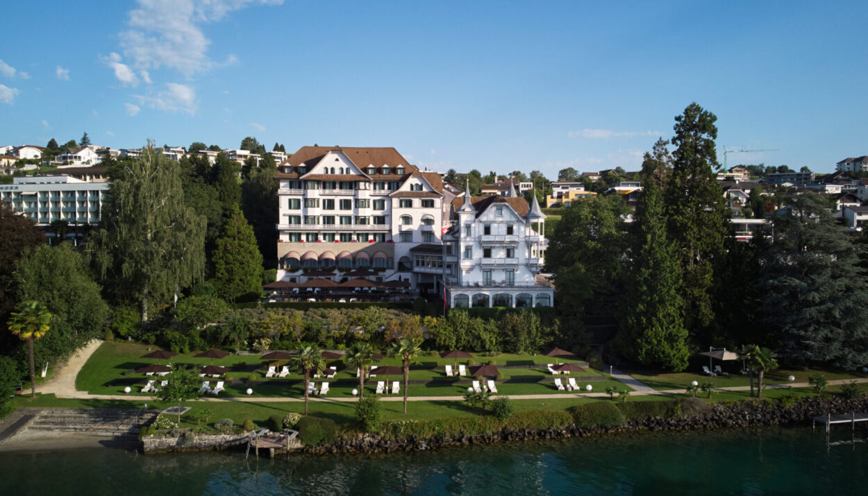 Veduta del Chenot Palace Weggis, un lussuoso resort sul lago di Lucerna circondato da giardini curati, con sdraio e ombrelloni che si affacciano sull'acqua, immerso in un ambiente naturale tranquillo e rilassante.