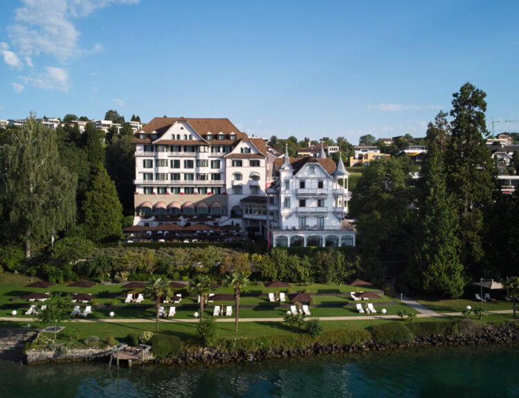 Veduta del Chenot Palace Weggis, un lussuoso resort sul lago di Lucerna circondato da giardini curati, con sdraio e ombrelloni che si affacciano sull'acqua, immerso in un ambiente naturale tranquillo e rilassante.