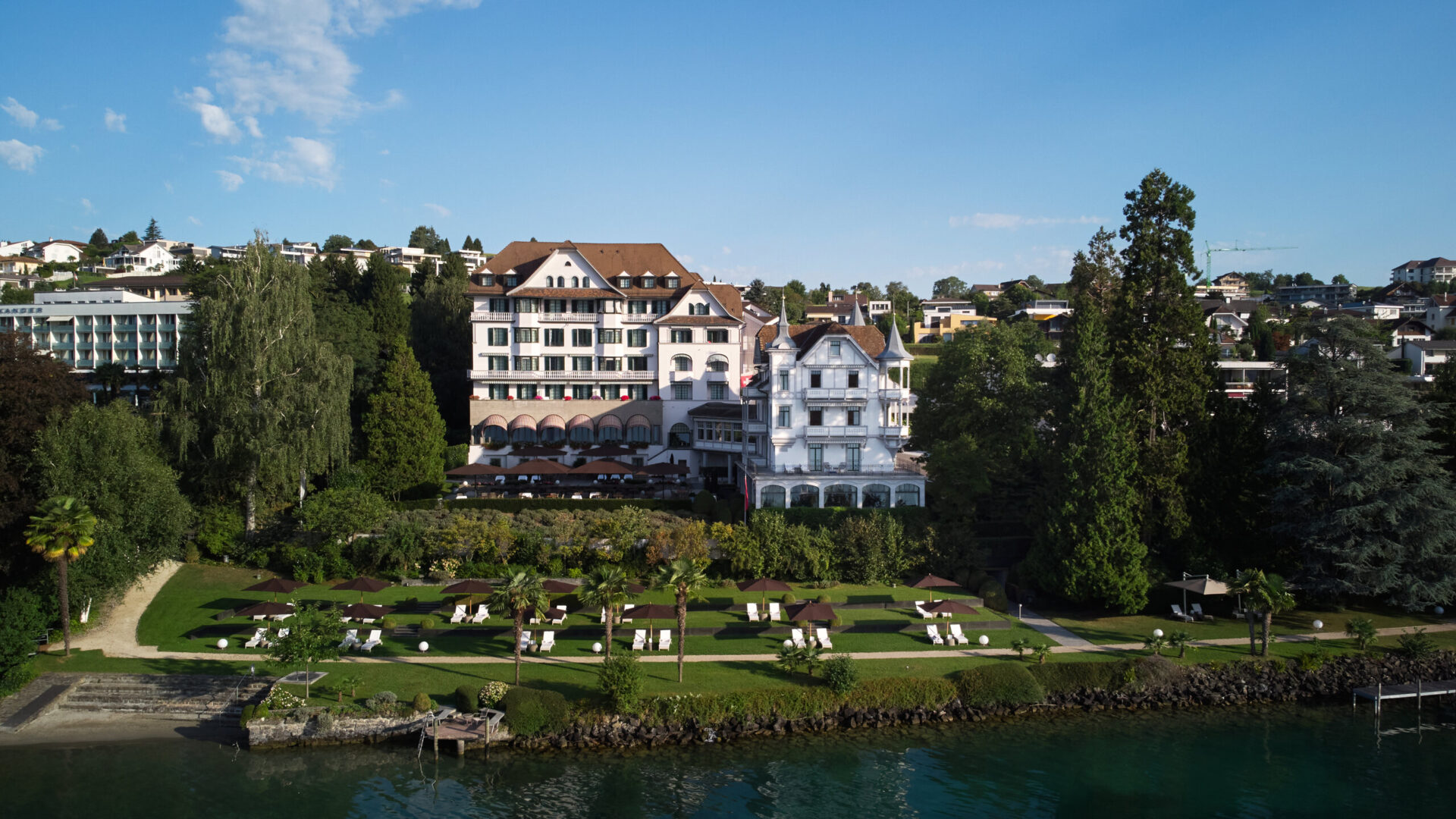 Veduta del Chenot Palace Weggis, un lussuoso resort sul lago di Lucerna circondato da giardini curati, con sdraio e ombrelloni che si affacciano sull'acqua, immerso in un ambiente naturale tranquillo e rilassante.