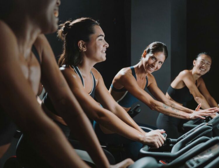 Un gruppo di donne sorridenti durante una sessione di indoor cycling, parte della Oysho Community, che mostra energia e spirito di squadra in palestra