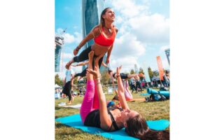 Due donne che praticano acroyoga in un parco durante l'evento Wanderlust 108 a Milano, con grattacieli sullo sfondo e altre persone in attività