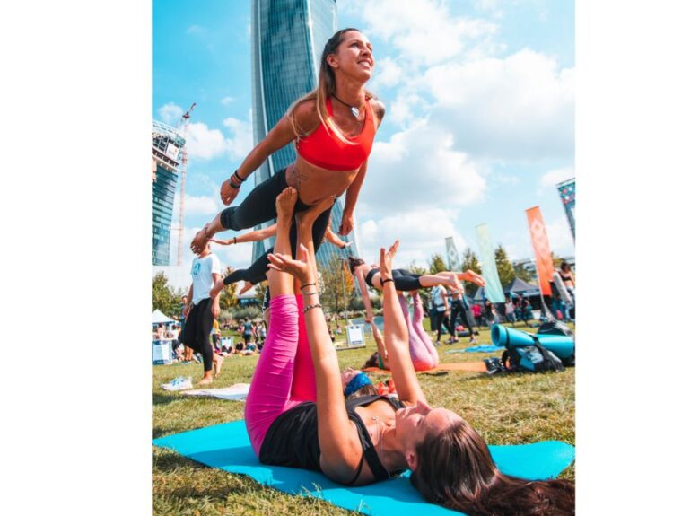 Due donne che praticano acroyoga in un parco durante l'evento Wanderlust 108 a Milano, con grattacieli sullo sfondo e altre persone in attività