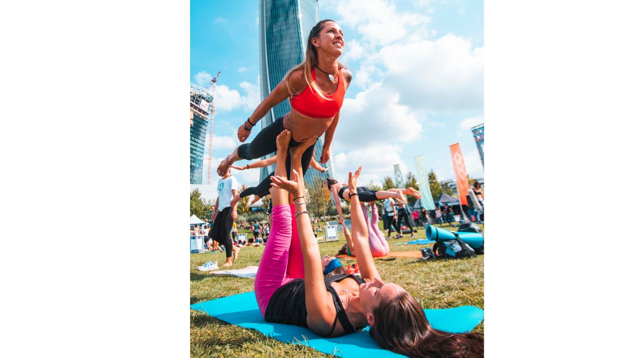 Due donne che praticano acroyoga in un parco durante l'evento Wanderlust 108 a Milano, con grattacieli sullo sfondo e altre persone in attività